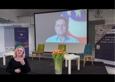 Zdjęcie kolorowe. Na scenie ekran ze zdalnym wystąpieniem pana Radosława Jasińskiego z Polsko-Amerykańskiej Fundacji Wolności, fundatora programu Fimango. W lewym dolnym rogu widoczna tłumaczka języka migowego