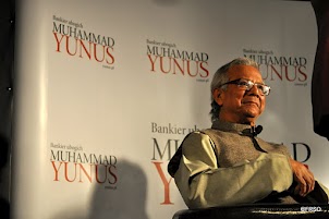 Plan półpełny. Siedzący Profesor Muhammad Yunus, osoba starsza z siwymi włosami w okularach. Na sobie ma koszulę i beżowy bezrękawnik. W tle plansza z napisami Bankier ubogich Muhammad Yunus.