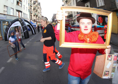 Na pierwszym planie człowiek przebrany za klauna w czerwonej koszuli, z pomarańczową dużą kokardą i brązowym kapeluszem, pozuje z drewnianą złotą ramką. W tle na uliczce między budynkami stragany i ludzie.